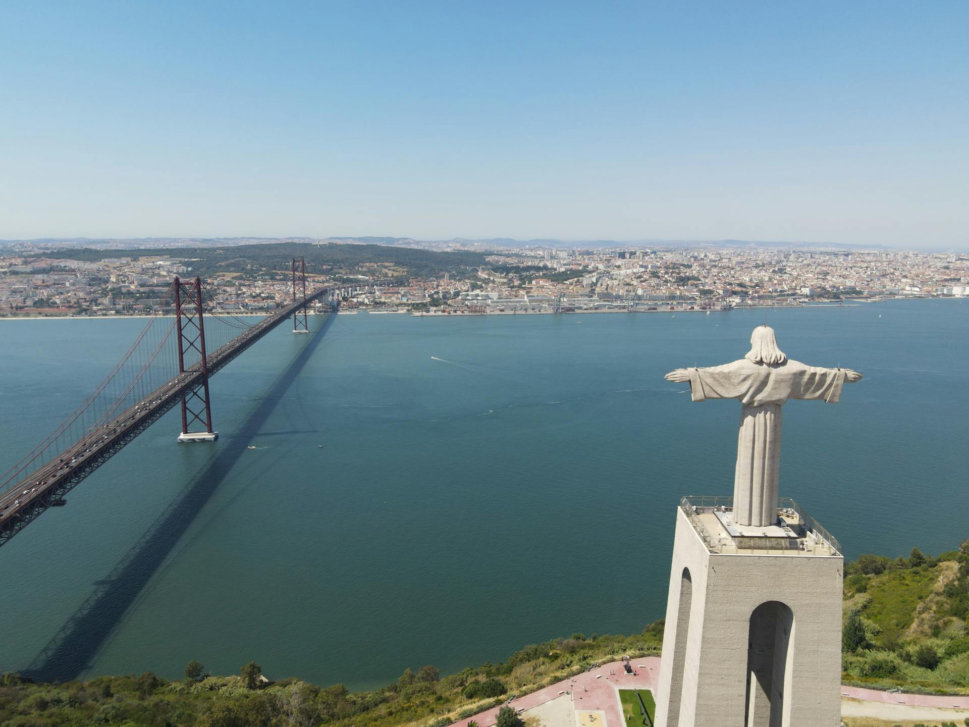 A striking aerial shot of the Christo Rei statue with the 25 de Abril Bridge and Lisbon cityscape in the background.