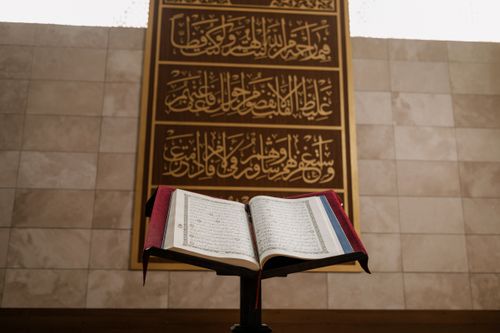 Free An open Quran on a wooden stand in front of ornate Islamic calligraphy inside a mosque. Stock Photo
