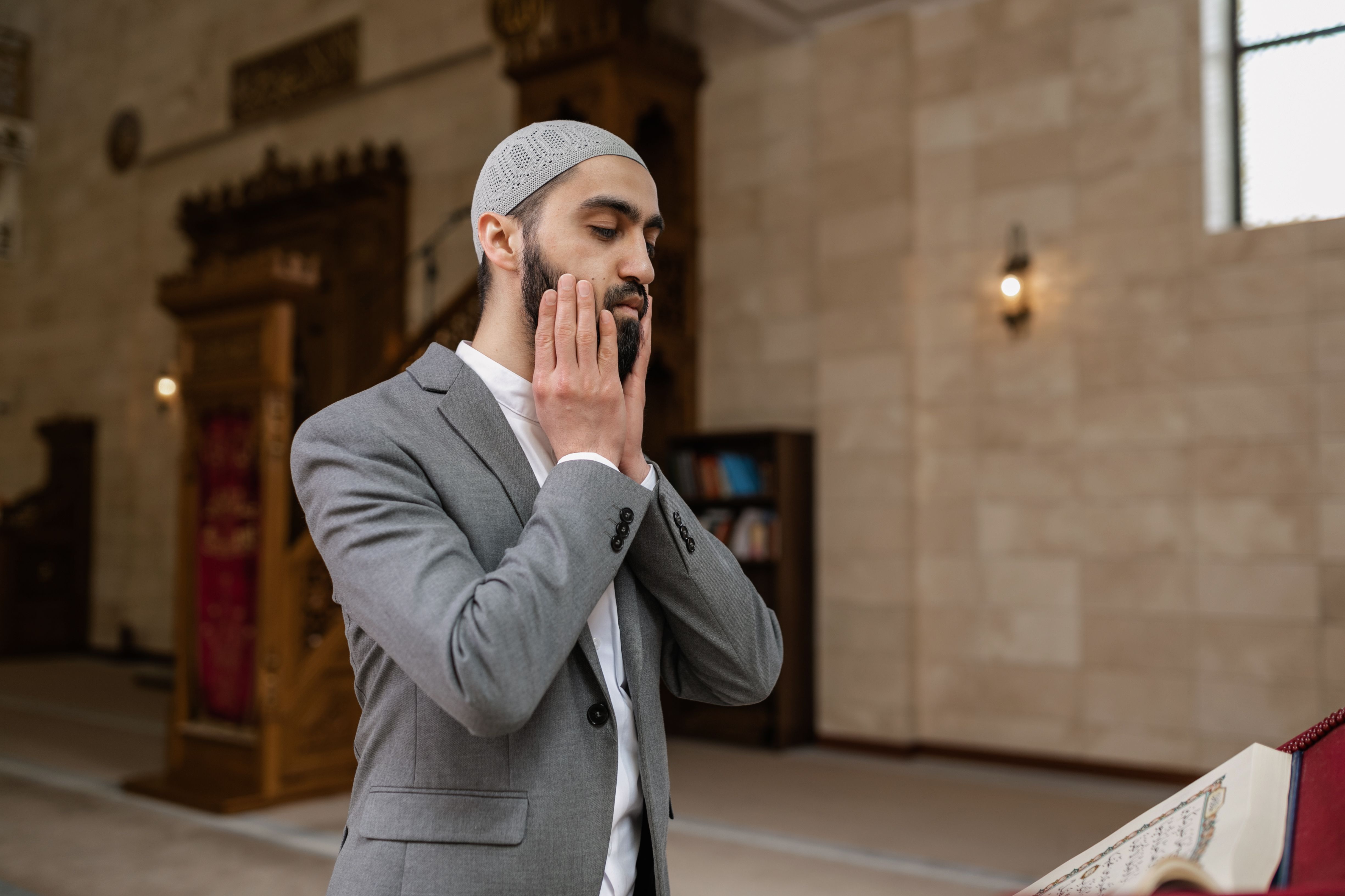 Asian man in a gray suit praying inside a mosque, showcasing Islamic culture and tradition.