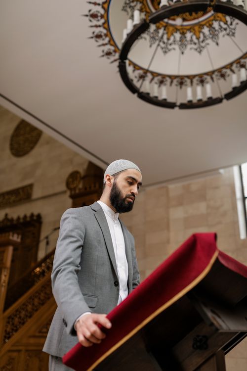 Free A man in a grey suit reads at a lectern inside a mosque, capturing a moment of reflection. Stock Photo
