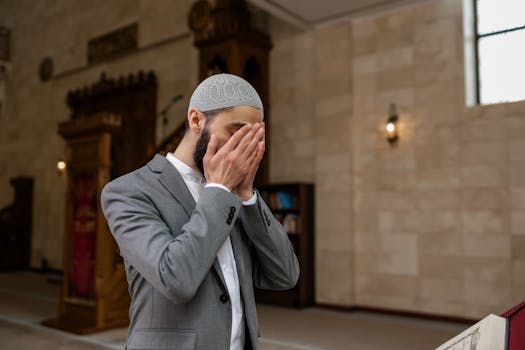 Middle Eastern man in a gray suit praying inside a mosque, covering his face with hands.