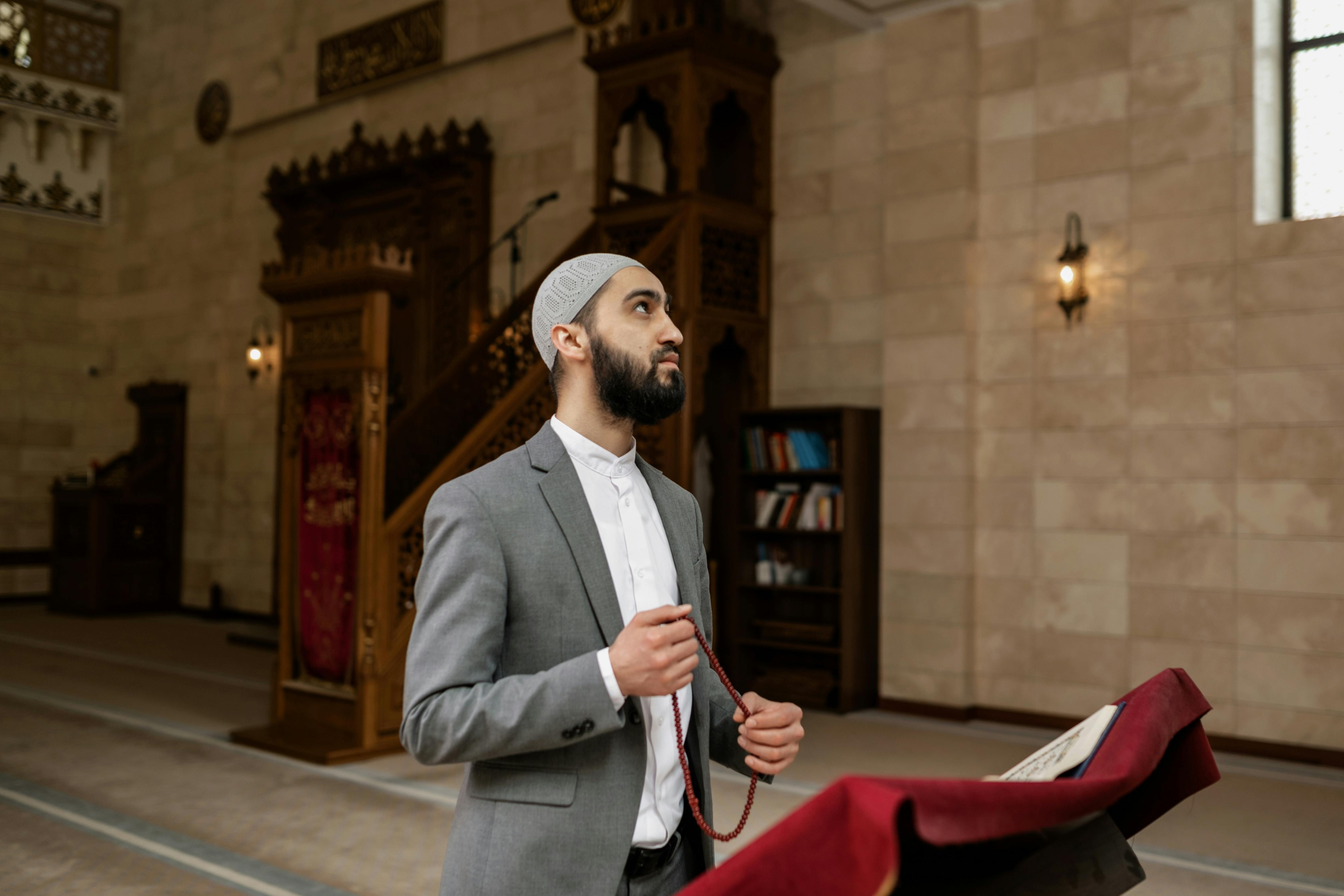 Man wearing a suit and skullcap holds prayer beads in an ornate mosque interior during Ramadan.