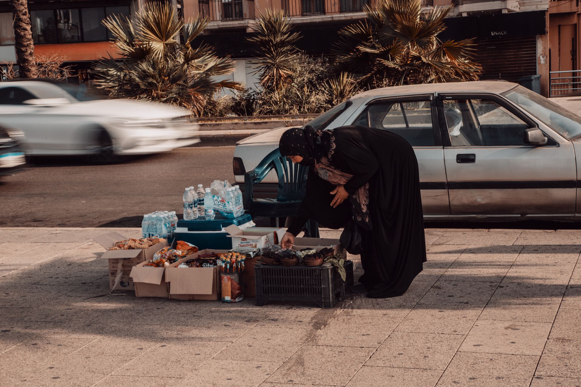 A woman vendor arranging snacks for sale on a busy city street with passing cars.