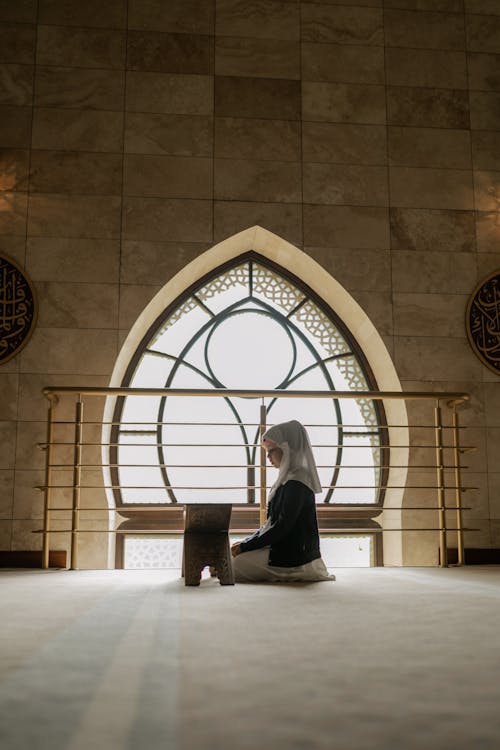 Woman in White Hijab Sitting on the Floor in Front of a Book Stand Backlit by Daylight from the Arch Window