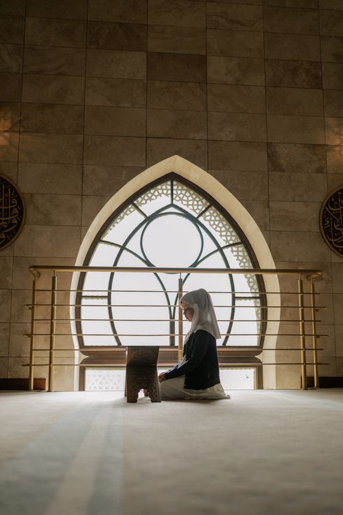 Free A young Muslim woman wearing a hijab sits praying with a Quran stand in a beautifully lit mosque. Stock Photo