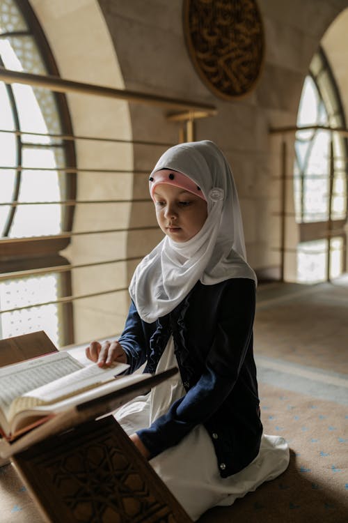 Girl in White Hijab and Blue Long Sleeve Shirt Sitting on Floor in Front of a Book