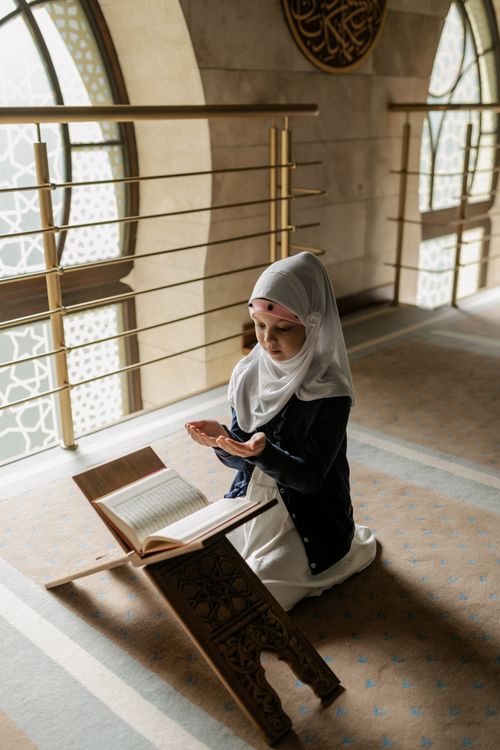 Free A young Muslim girl in a hijab prays indoors beside an open Quran on a stand. Stock Photo