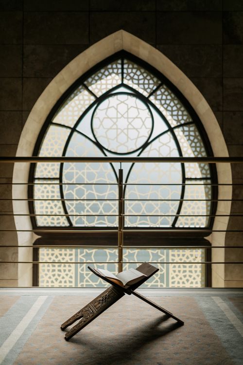 Free Open Quran on a wooden stand in front of an ornate arched window inside a mosque. Stock Photo
