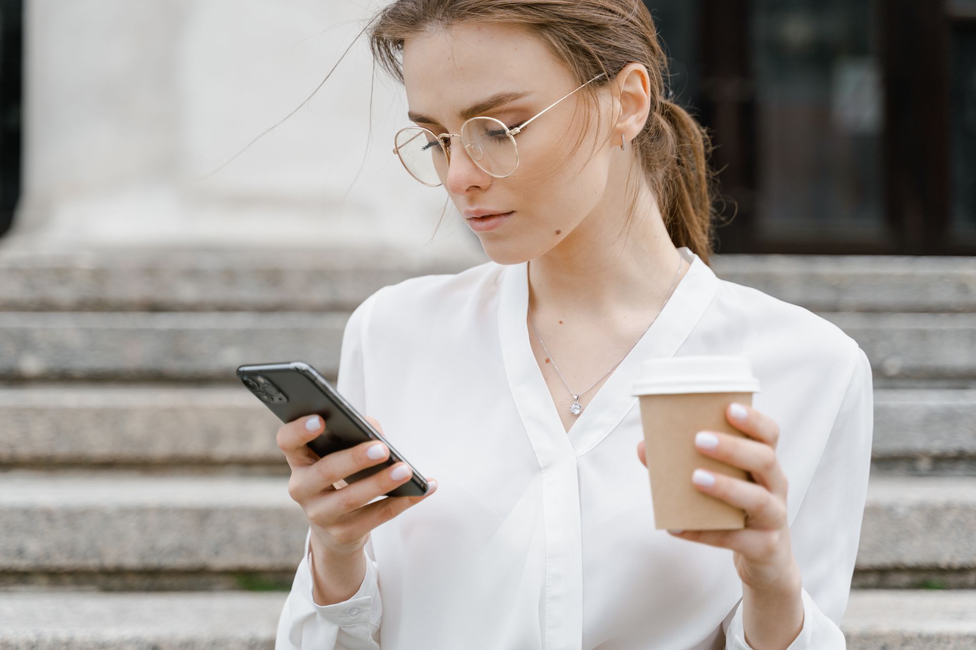 Young woman in white shirt browsing smartphone while holding coffee outdoors.