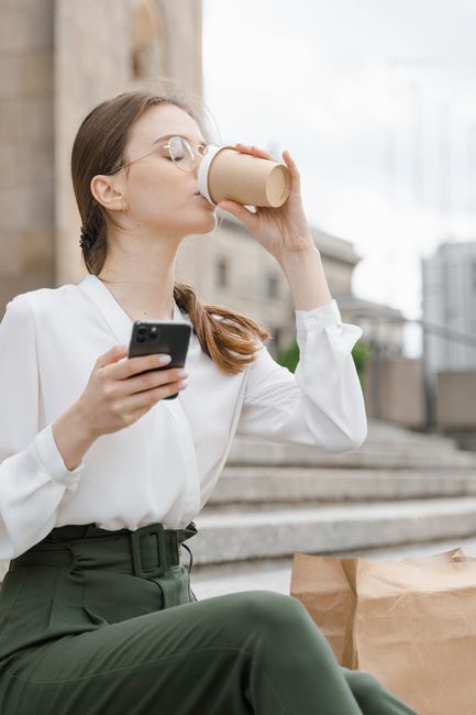 Woman enjoying coffee and using smartphone outdoors on a sunny day.