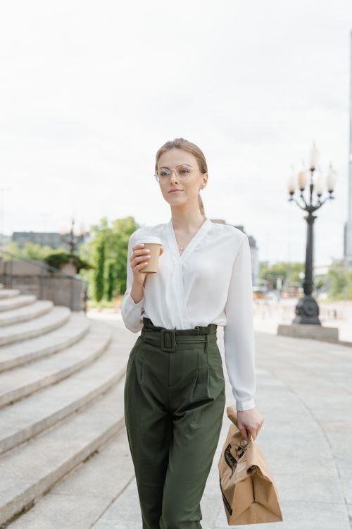 Free Confident woman in white blouse holding coffee and paper bag while walking outside on a sunny day. Stock Photo