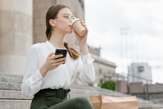 Young woman sipping takeaway coffee and using smartphone on outdoor steps.