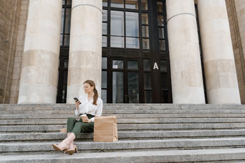 
A Woman Sitting on Stairs while Having Coffee and Using Her Smartphone