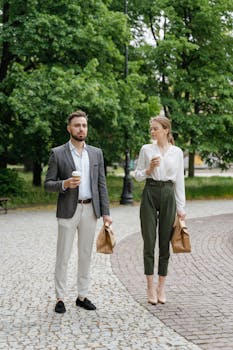 A man and woman stroll through a park with coffee and lunch bags in hand.