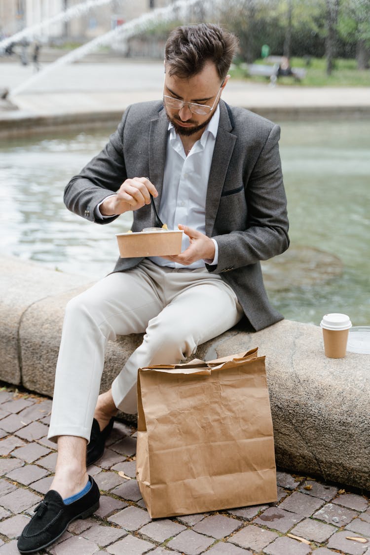 Man Wearing Gray Jacket Sitting By The Water Pond Eating Takeaway Meal