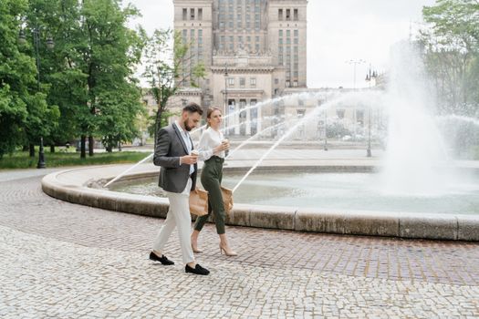 Professional man and woman walking near a city fountain, carrying coffee and takeout in a pleasant urban setting.