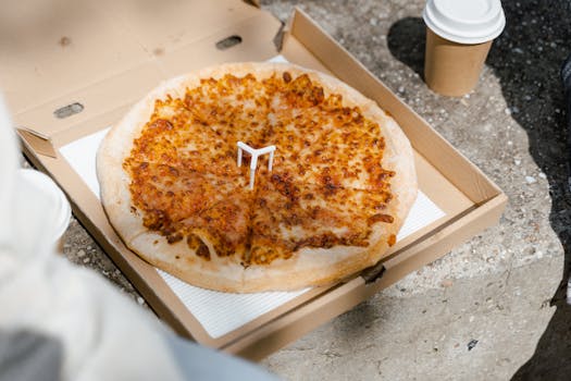 Cheesy pizza in an open box with a disposable cup, perfect for a casual outdoor meal.