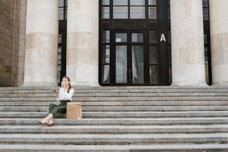 Woman Sitting On A Concrete Stairs At The Office Entrance Drinking Coffee And Using Handphone