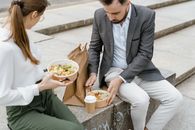 Man and woman enjoying takeaway lunch on stairs in a park. Casual outdoor meal with hot beverage and paper bags.