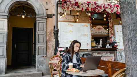 A woman at an outdoor cafe working on a laptop with coffee and snacks.