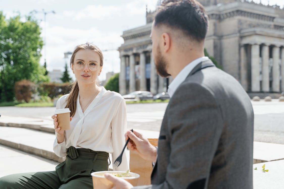 A female and a male coworker sit on the steps outside for a break while she takes a drink, and he eats his lunch.
