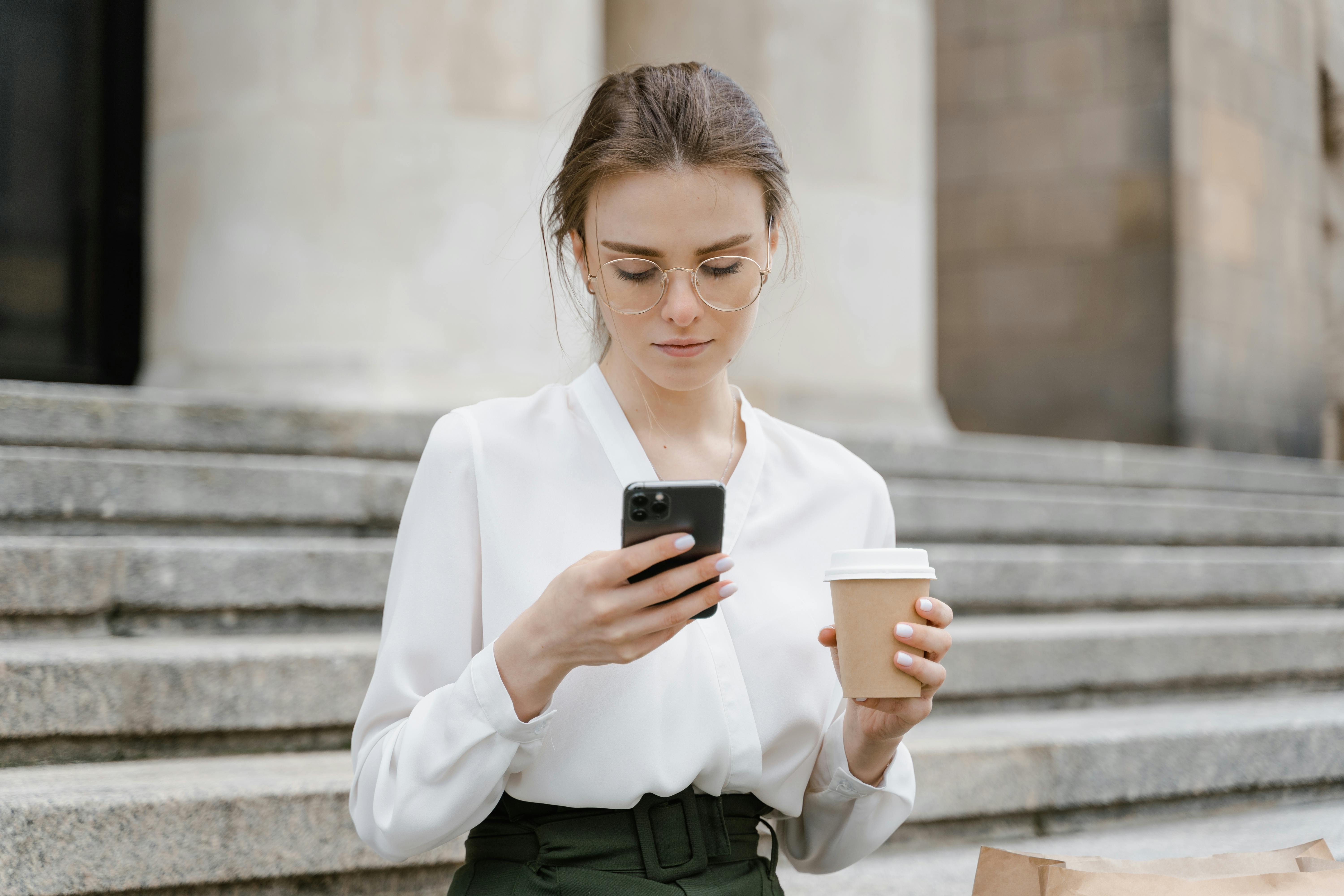 A young woman in glasses texting on her phone while holding a coffee cup outdoors.
