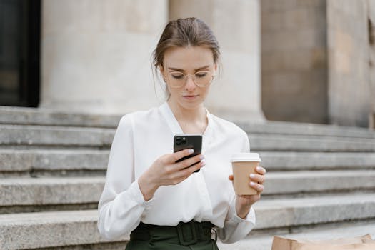 A young woman in glasses texting on her phone while holding a coffee cup outdoors.