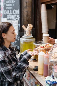Woman in plaid shirt buying waffles at a street food stall with sauces and drinks visible.