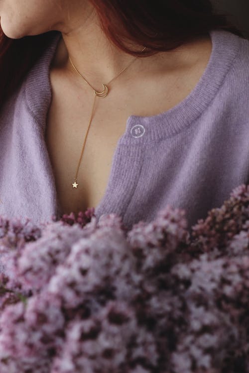 Crop faceless female with pendant in purple soft sweater and bouquet of blossoming flowers in hands standing in light room