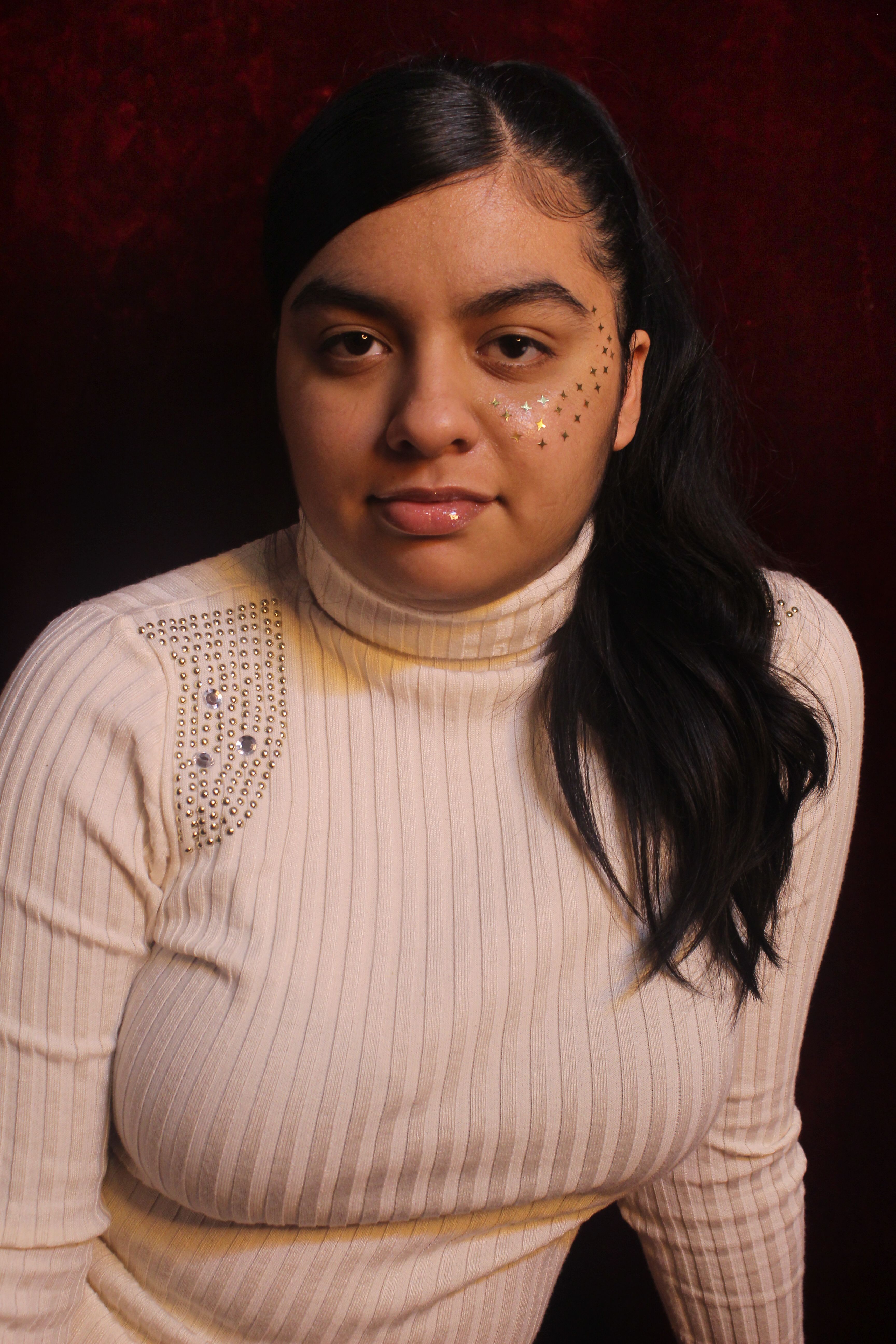 Young woman wearing a white turtleneck and face stickers, posing in a warm indoor setting.