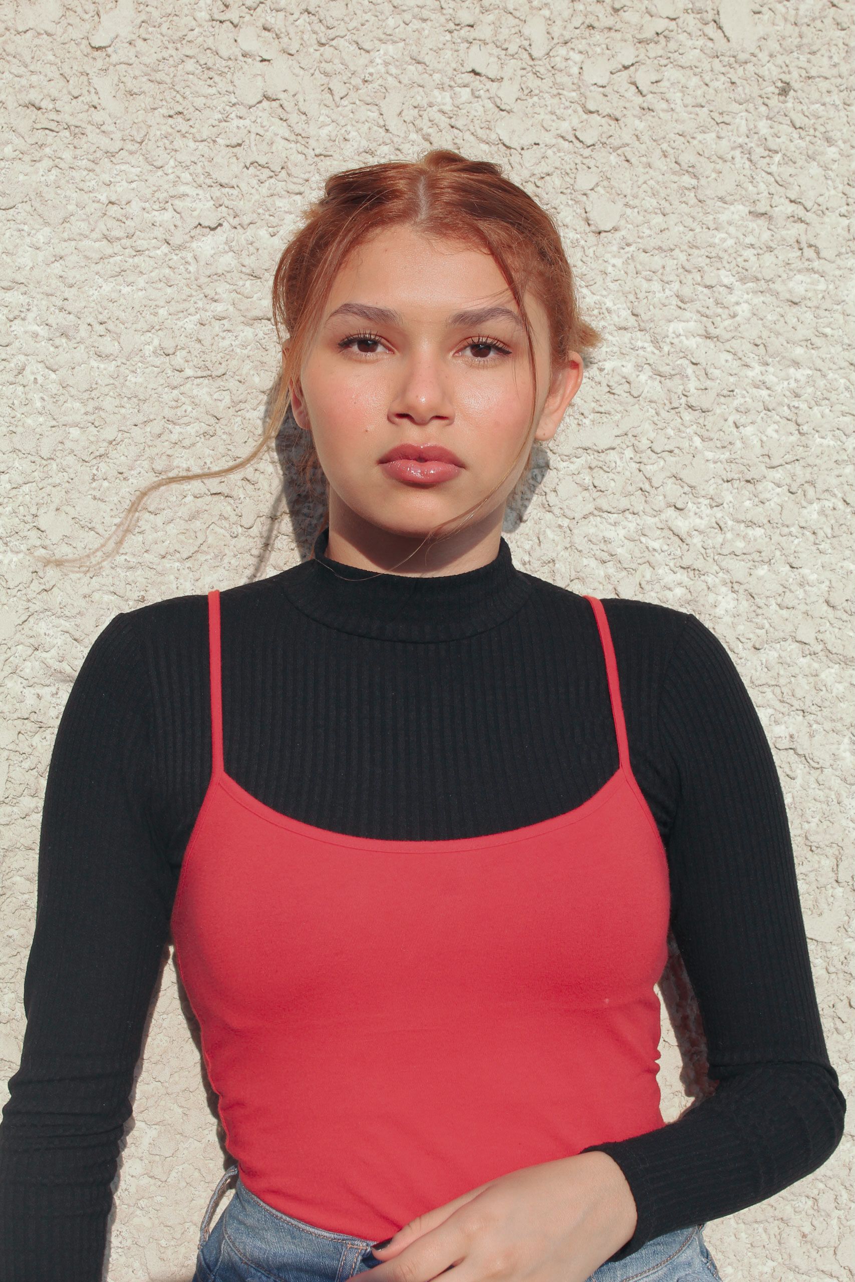Young woman in red tank top and black sweater against a textured wall, basking in sunlight.