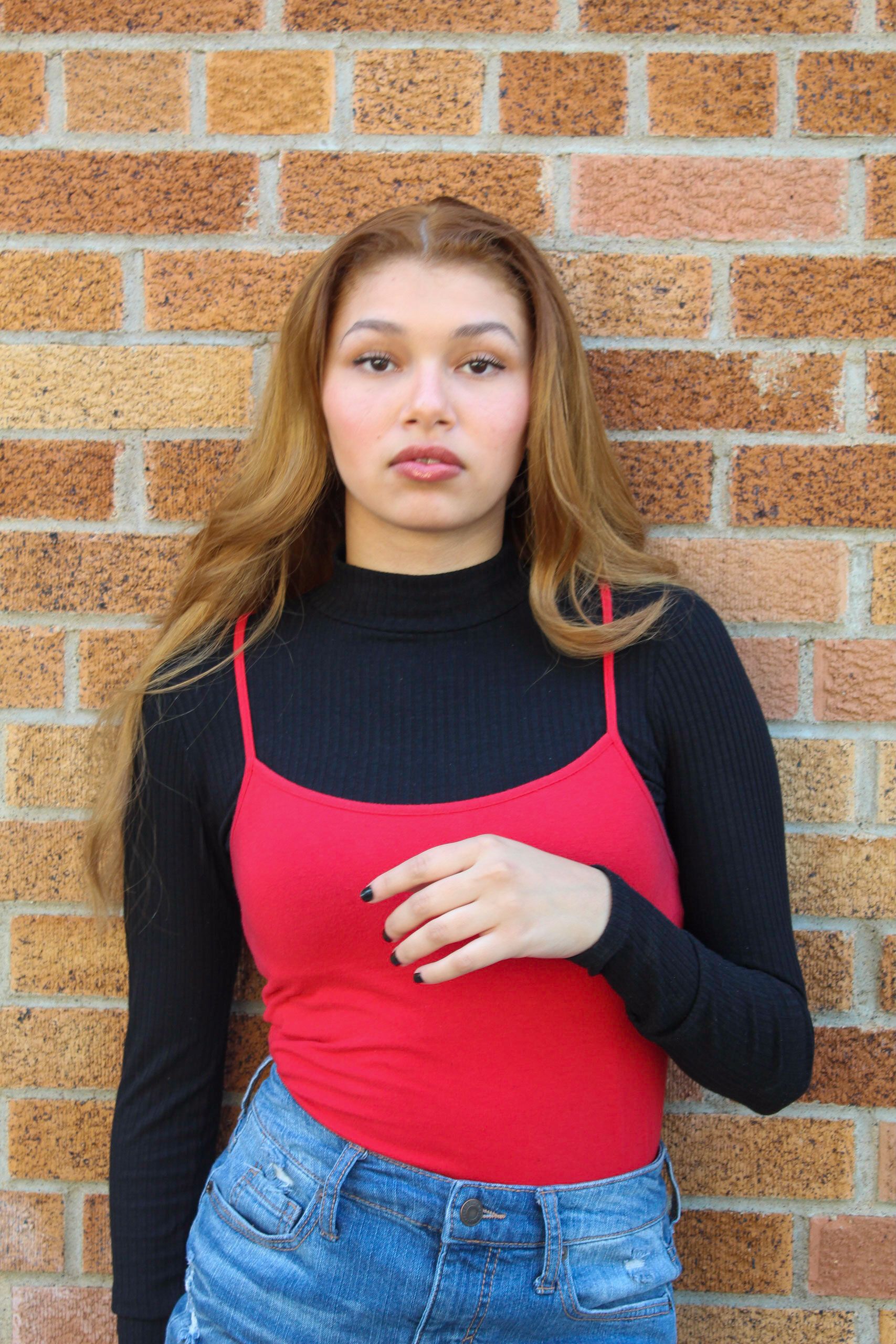 A fashionable woman in a red top and jeans posing confidently against a brick wall.