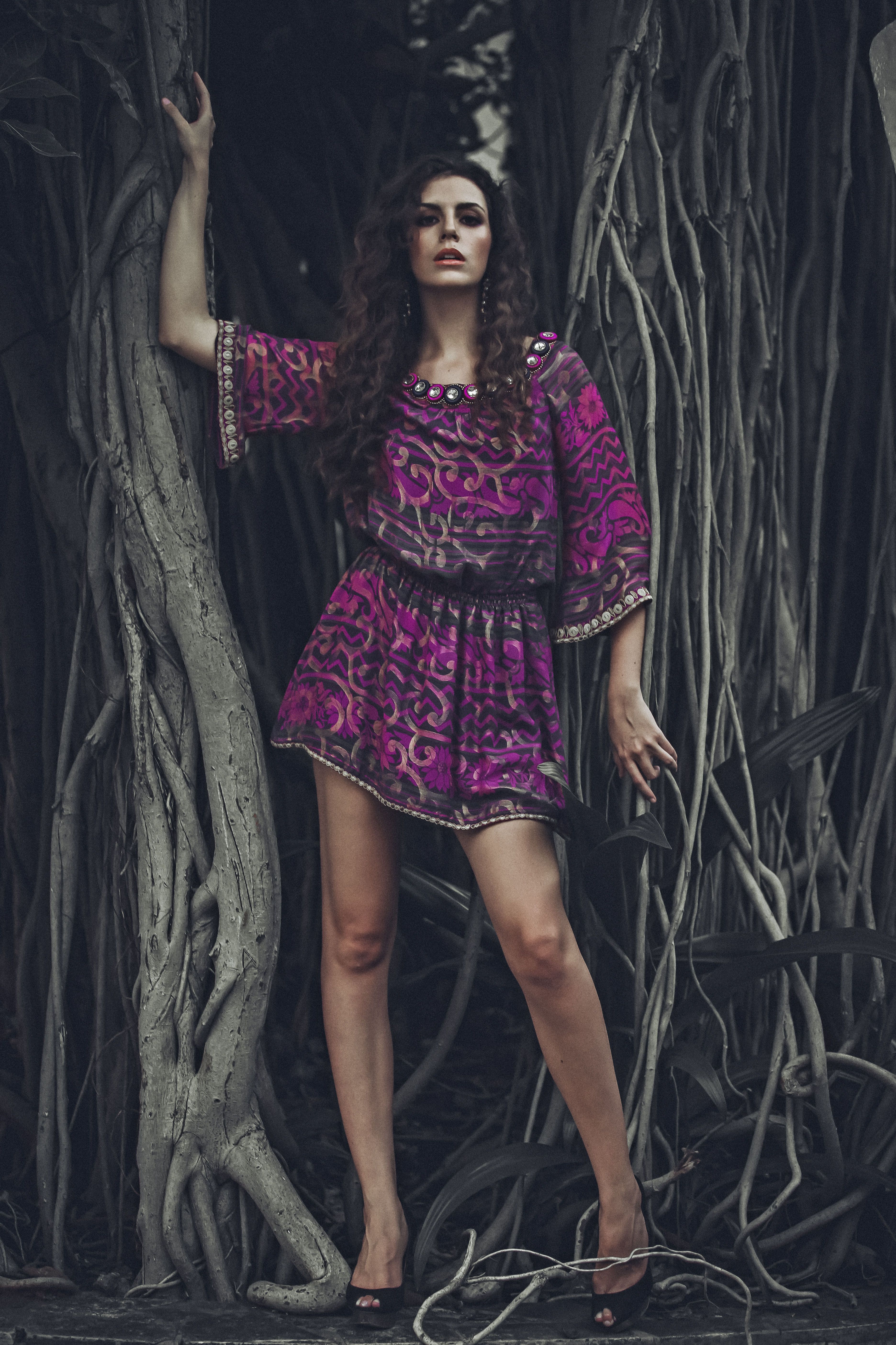 Elegant woman in a purple dress poses among dramatic tree roots, showcasing fashion and nature.
