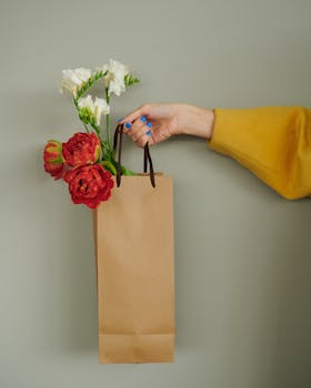 A stylish hand holding a bouquet in a paper bag against a neutral green backdrop. Perfect for gift themes.