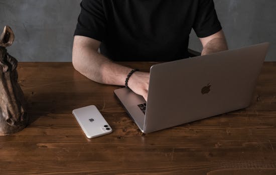 From above of crop unrecognizable male in black shirt sitting at wooden desk with laptop and smartphone while working