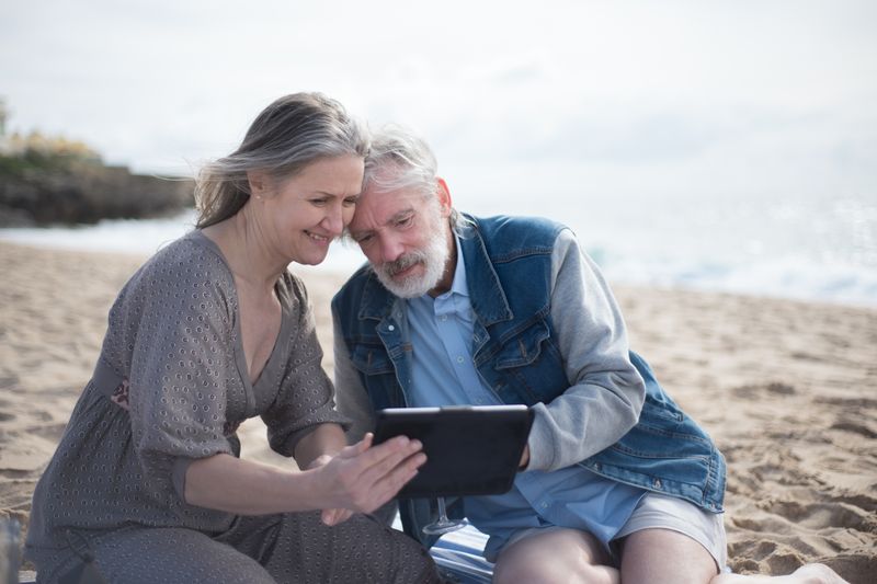 Senior couple sharing a joyful moment on a beach in Portugal, using a tablet.