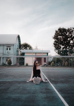 A stylish woman sitting on a tennis court in sunglasses, exuding a cool and relaxed vibe.