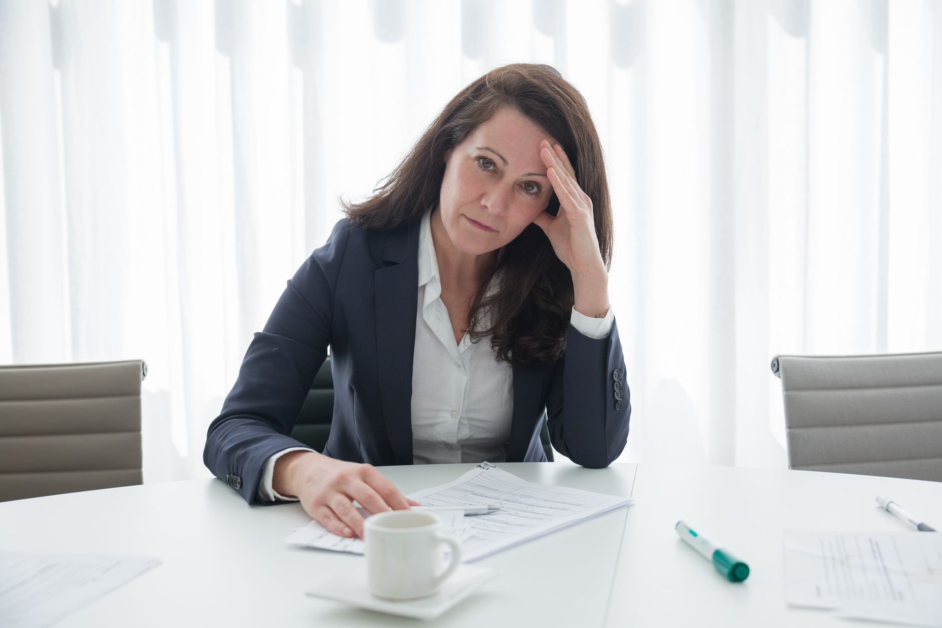 A stressed businesswoman in corporate attire sits at a desk with papers, looking overwhelmed.