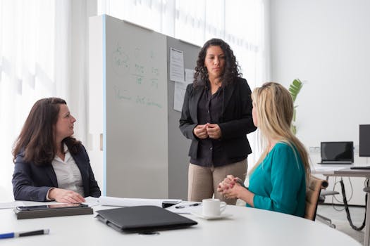 Three women in an office setting having a professional discussion, focused and engaged with a whiteboard.