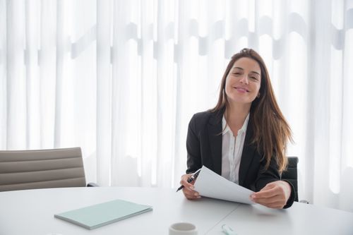Free Smiling businesswoman in a blazer holding paper at an office table indoors. Stock Photo