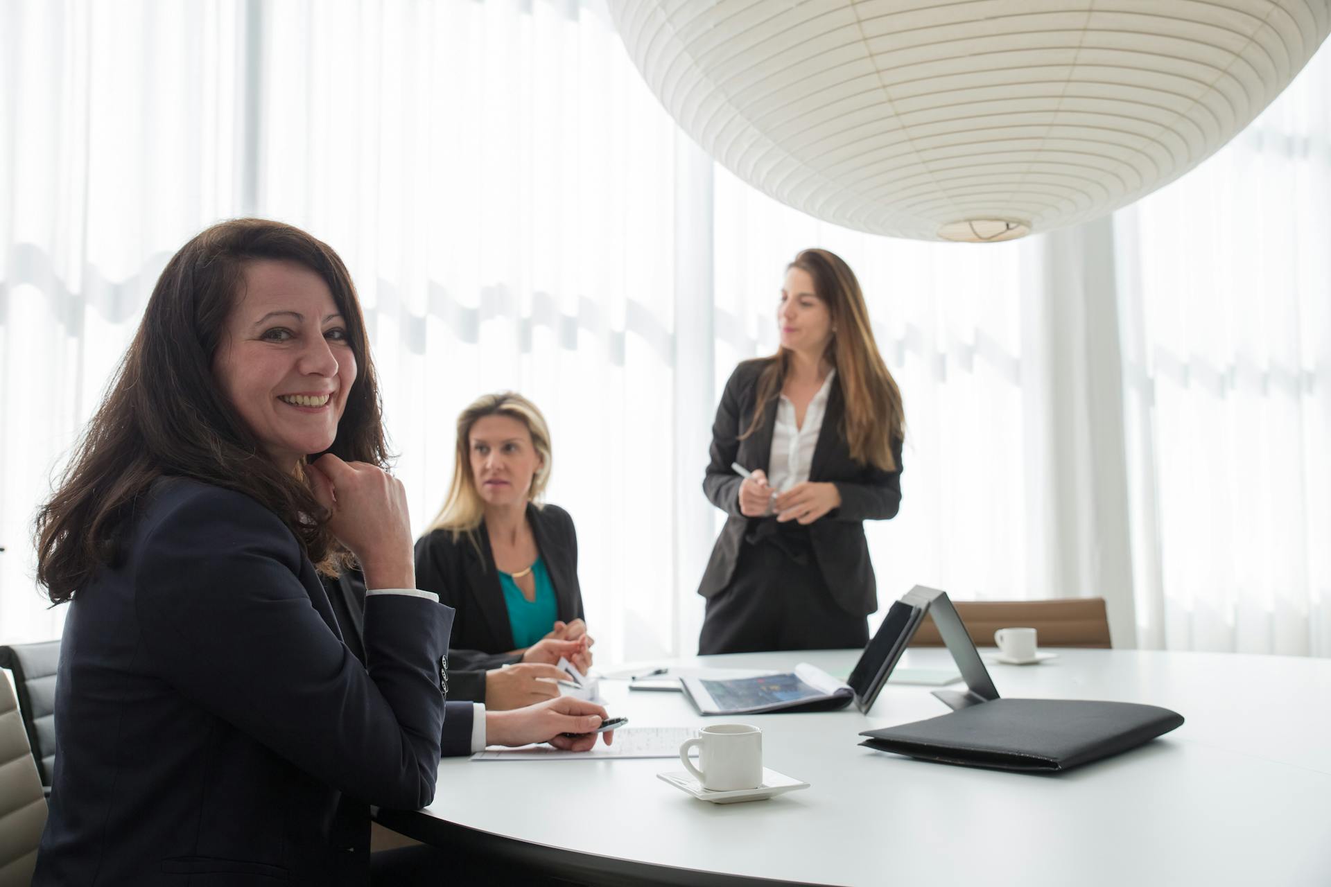 Three women in formal attire smiling and discussing business during a meeting.