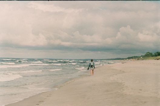 A person walking alone on a sandy beach with waves under a cloudy sky.