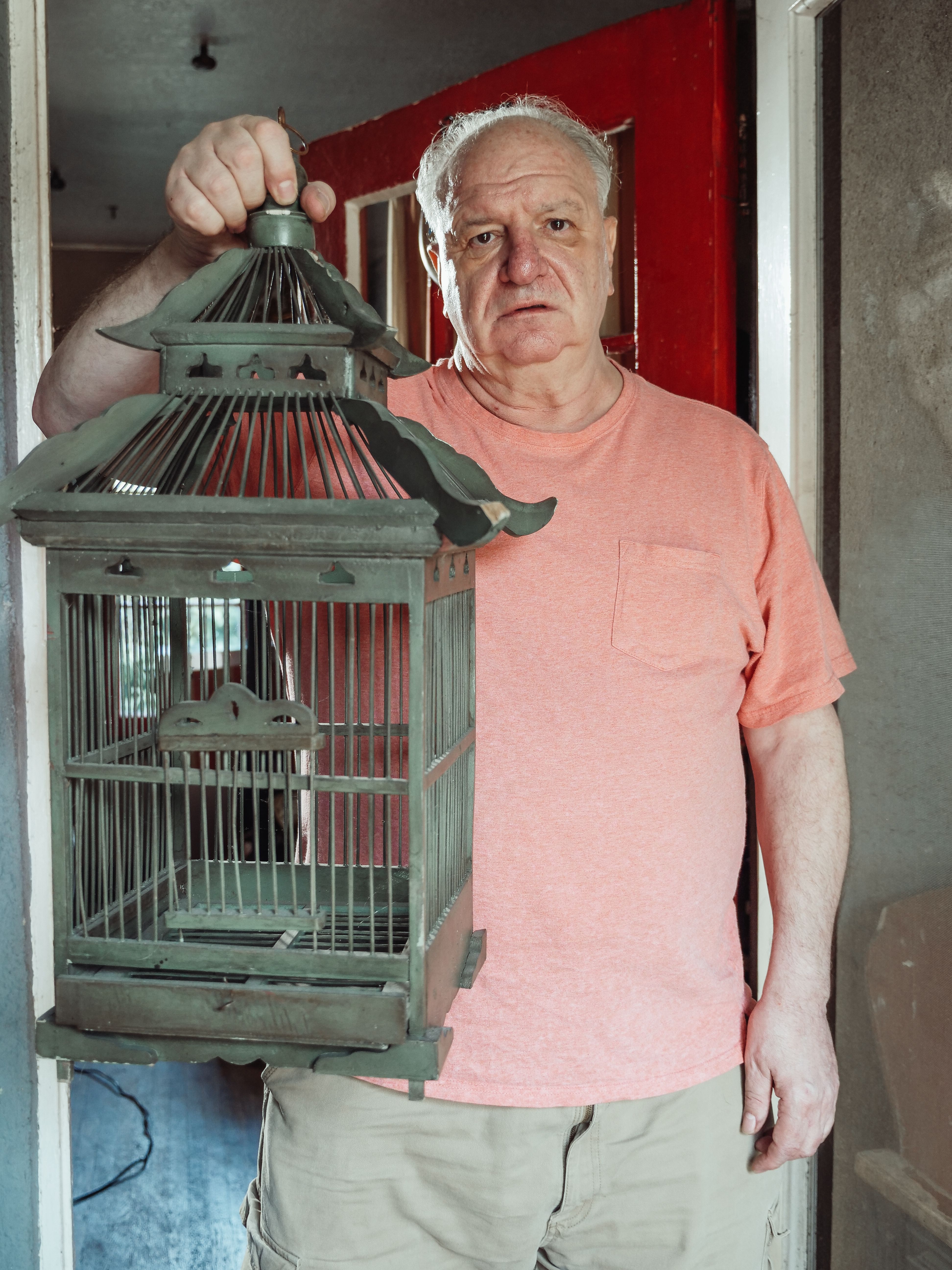 Elderly man in casual attire holding a vintage birdcage, standing indoors with a red door backdrop.