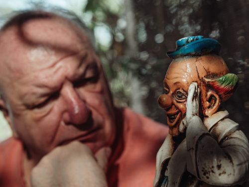 Free Elderly man in deep thought beside a colorful porcelain clown statue. Stock Photo