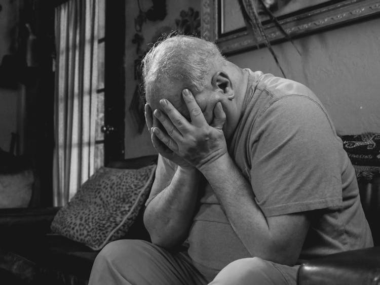 An Elderly Man Sitting On The Couch While Covering His Face