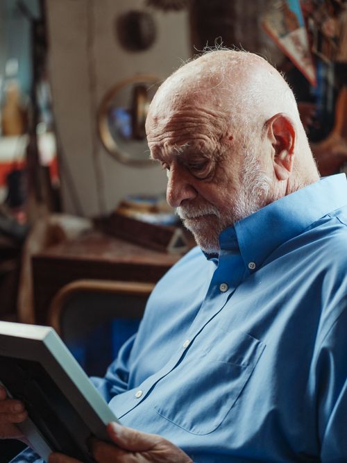 Free Senior man in a blue shirt sitting indoors, holding a picture frame, experiencing deep emotions. Stock Photo