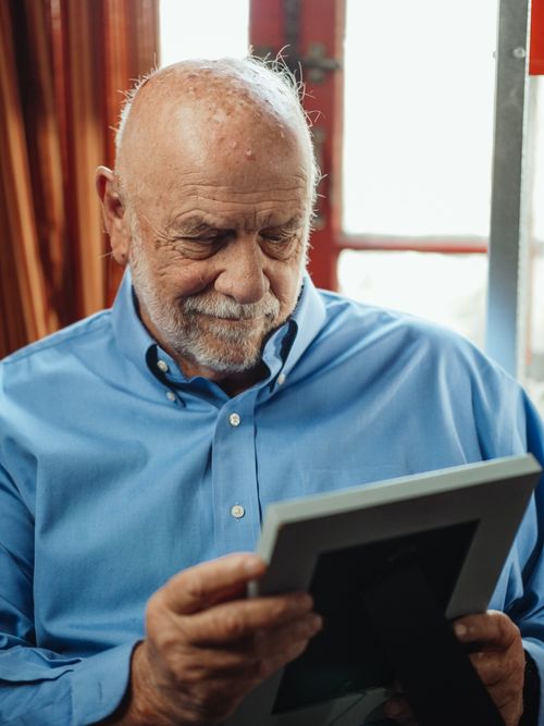 Free Senior man in blue shirt holding a photo frame, reflecting thoughtfully indoors. Stock Photo