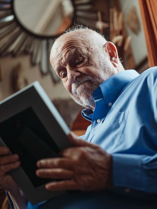 Free Elderly man wearing blue long sleeves emotionally holding a photo frame indoors. Stock Photo