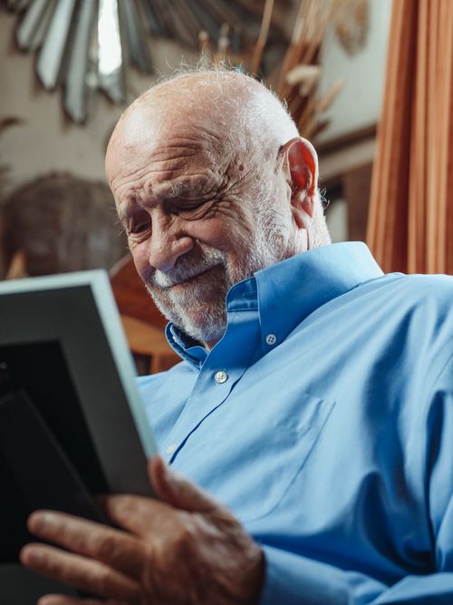 Free Elderly man in blue shirt holds photo frame, expressing grief indoors. Stock Photo