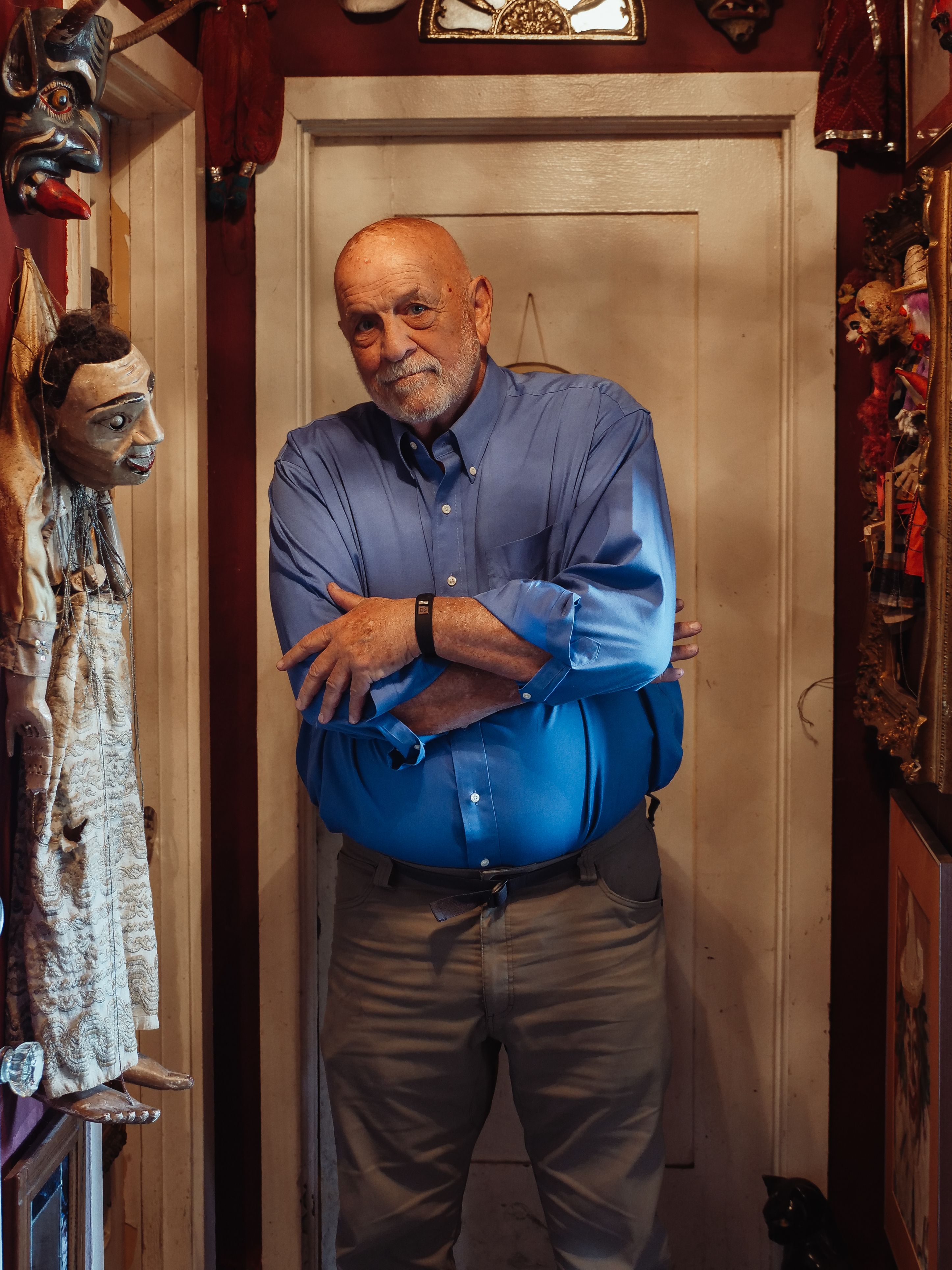 Senior man in blue shirt stands with arms crossed in vintage puppet-filled room.